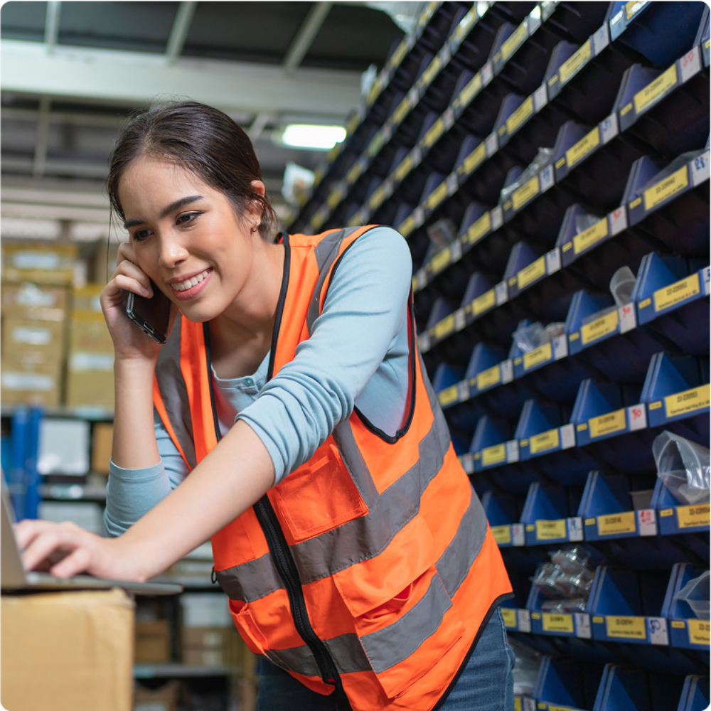 This image features a woman wearing a safety vest, working in a warehouse setting. She is multitasking, using a laptop and speaking on a mobile phone, surrounded by organized shelving filled with various items.