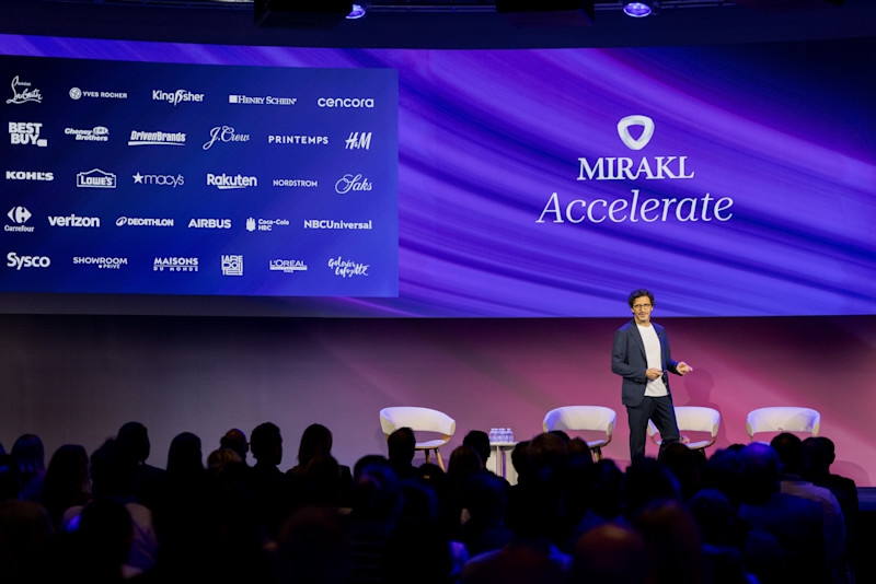 A man in a navy suit jacket and glasses stands on a stage with a microphone. He is gesturing with his hands. Behind him, a large screen displays the Mirakl logo and the word "Accelerate" on the right. The left side of the screen is a collage of company logos including Best Buy, Kohls, Lowes, Macys, Rakuten, Nordstrom, Saks, Carrefour, and others. The audience is visible in the foreground, with their backs to the camera.