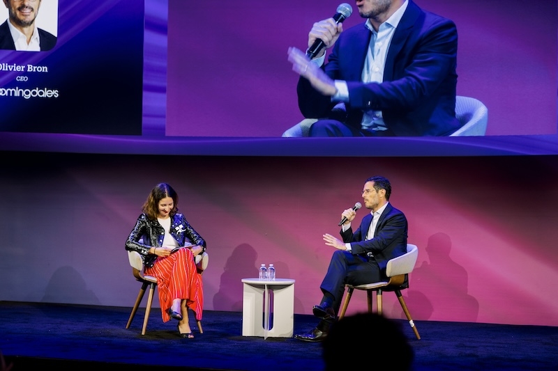 A man and a woman are seated on a stage, facing each other in conversation. The man on the right, in a dark suit, holds a microphone and gestures with his hands. The woman on the left is wearing a shiny black jacket and a long, red and white striped skirt. A small table with two bottles of water sits between them. A large screen behind them shows a close-up of the man's face and a graphic with the name Olivier Bron, his title, and the Bloomingdale's logo.