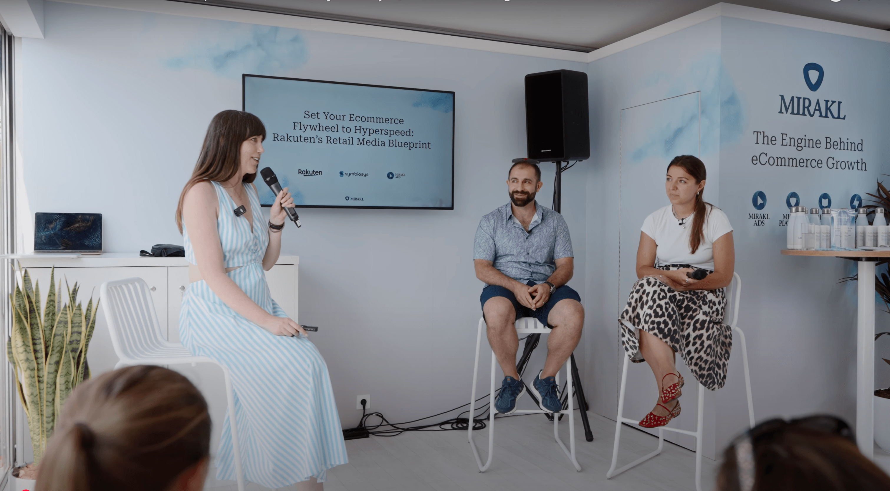 Three people are seated on high stools on a small stage during a Mirakl Mornings session at Cannes. A woman on the left, Anne Hallock, VP, Sales for Mirakl Ads, is speaking into a microphone. In the center, a man with a beard, Bashar Kachachi, Founder and CEO of Symbiosys, is seated. On the right, a woman, Kristina Raptovaia, Rakuten France's Head of Partnerships and Advertising, is seated. The large screen behind them displays the title "Set Your eCommerce Flywheel to Hyperspeed: Rakuten's Retail Media Blueprint," and the event discussed how their collaboration fuels a more connected, performant retail media ecosystem. The wall behind the stage features a cloud pattern and the "MIRAKL" logo.