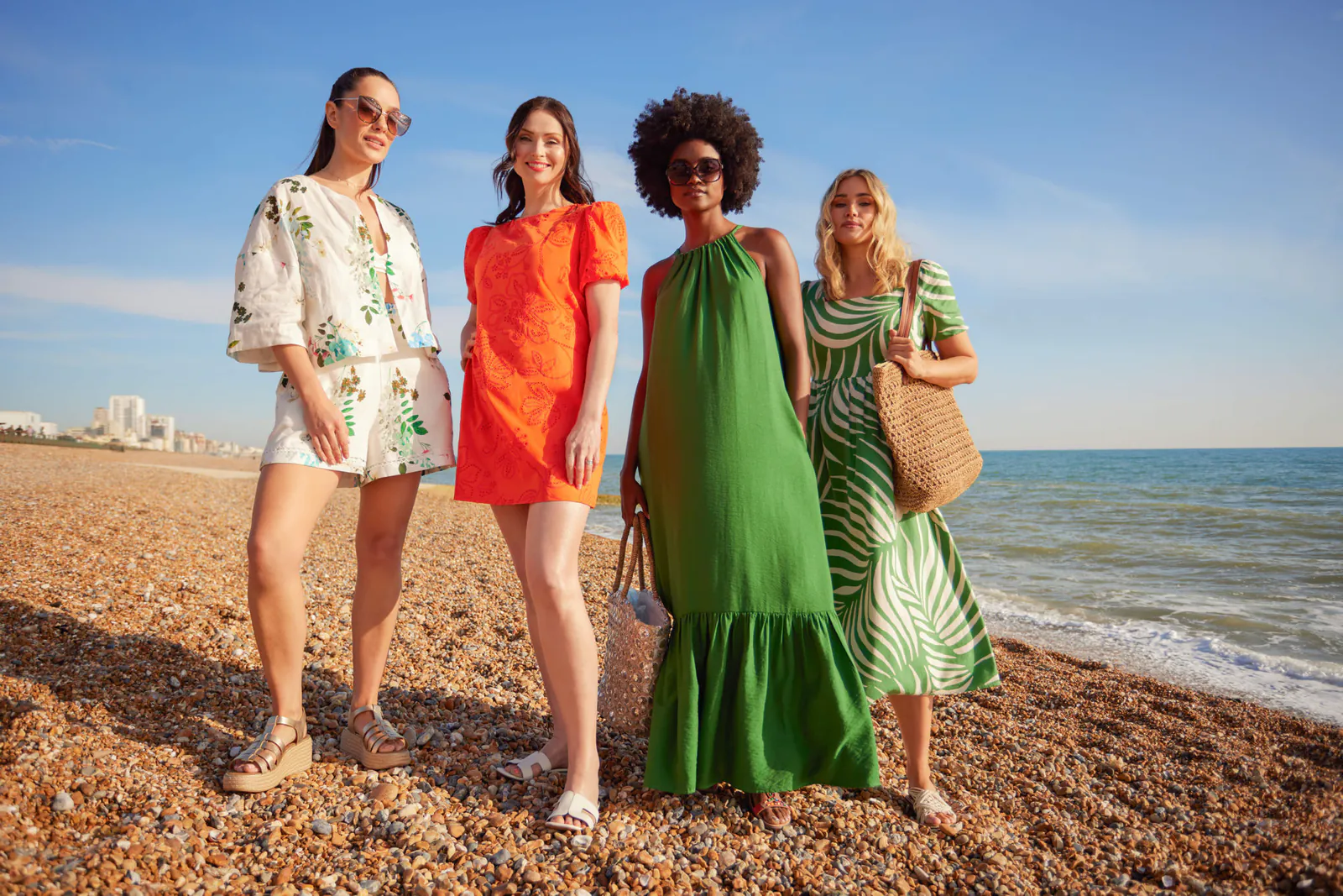 Four women in summer outfits stand on a sunlit pebble beach by the sea: a white floral linen co-ord, an orange broderie-anglaise dress, a green halter-neck maxi dress, and a green-and-white palm-print dress paired with a straw tote bag.
