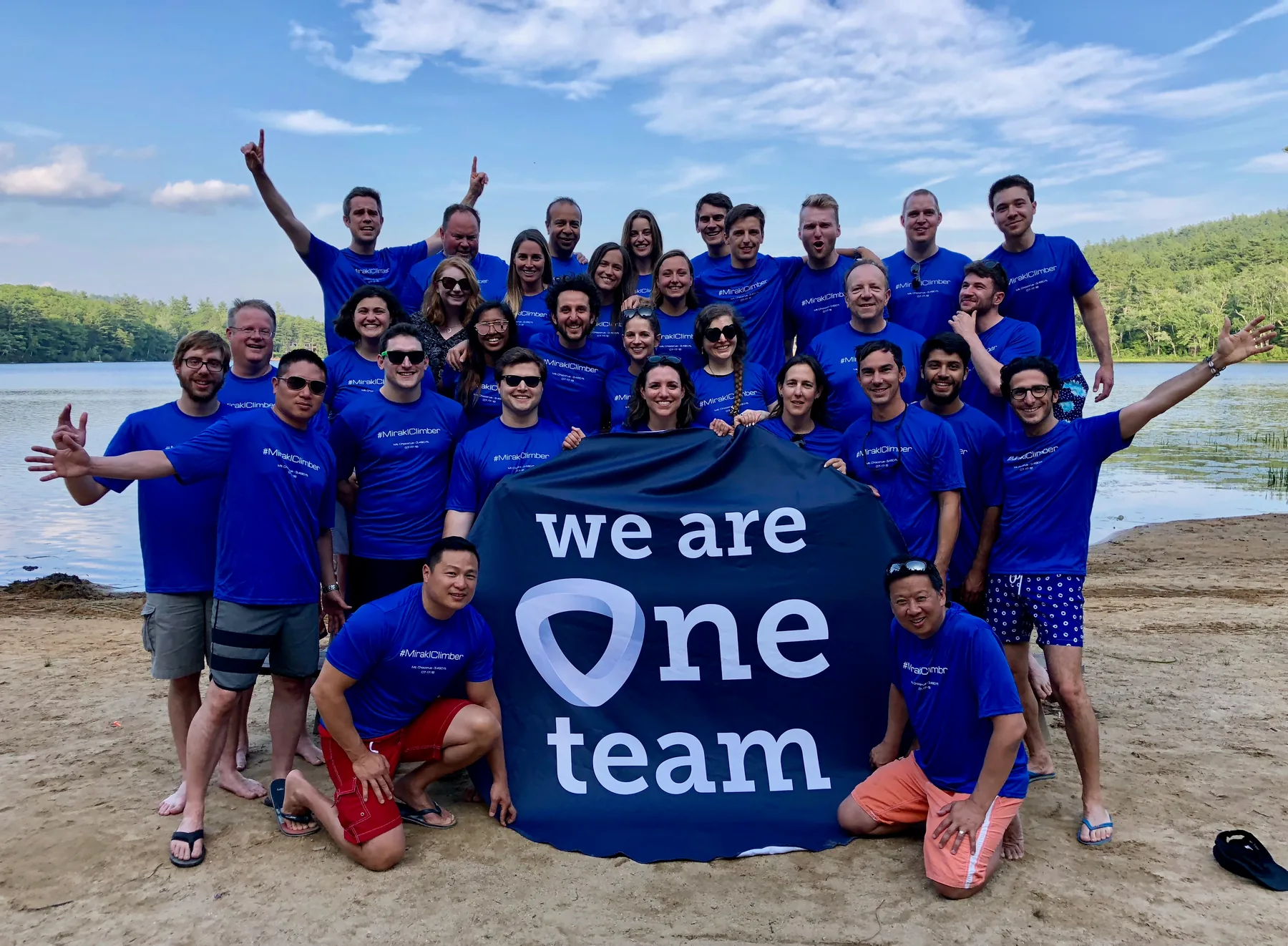 Group photo of approximately 25 Mirakl Boston team members posing outdoors on a sandy beach in matching blue t-shirts, holding a banner that reads "we are One team" with the Mirakl logo.